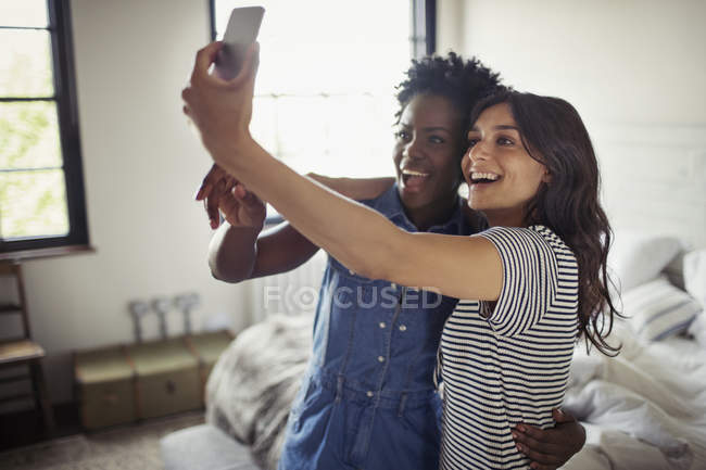 Улыбающаяся лесбийская пара обнимается, делает селфи с телефоном в спальне — стоковое фото