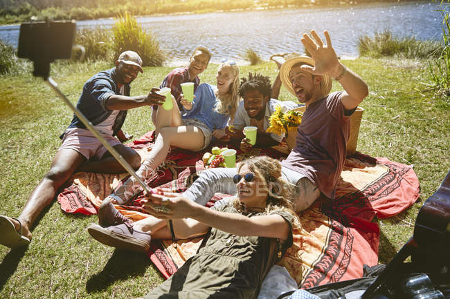 Грайливі молоді друзі беруть селфі з палицею, насолоджуючись пікніком на сонячному літньому березі річки — стокове фото