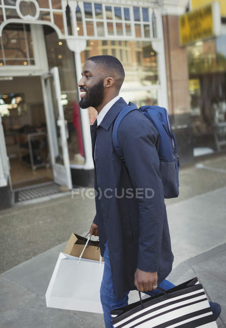 Улыбающийся молодой человек, идущий вдоль витрины магазина, несущий сумки — стоковое фото