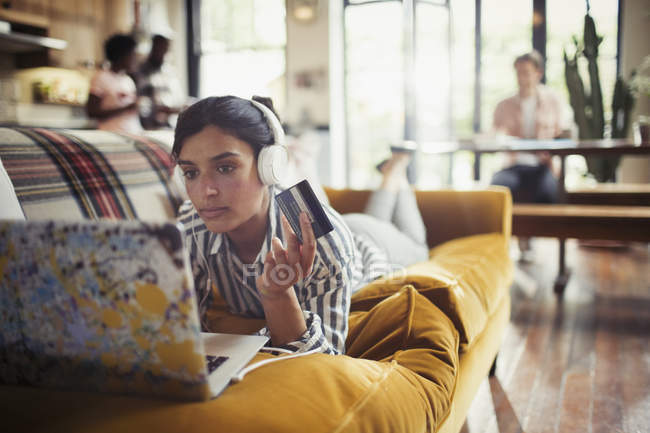 Молода жінка з навушниками та кредитною карткою онлайн покупки на ноутбуці на дивані у вітальні — стокове фото