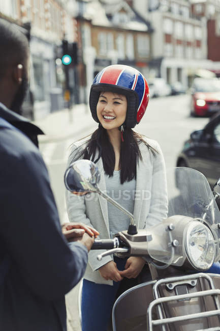 Lächelnde junge Frau mit Helm auf Motorroller, spricht mit Freund auf der Straße — Stockfoto