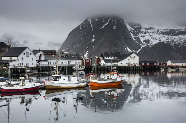 Barcos de pesca e aldeia à beira-mar abaixo nevado, montanhas acidentadas, Hamnoya, Lofoten, Noruega — Fotografia de Stock