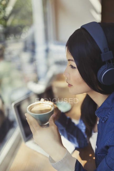 Pensive young woman drinking coffee, listening to music with headphones at cafe window — Stock Photo
