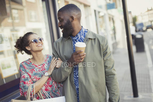 Sorridente giovane coppia con caffè a piedi braccio in braccio lungo vetrine — Foto stock