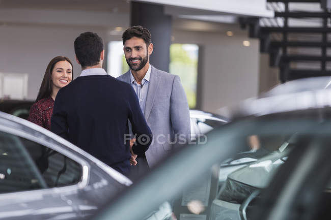 Vendedor de coches saludo, estrechando la mano con un par de clientes en la sala de exposición concesionario de automóviles - foto de stock