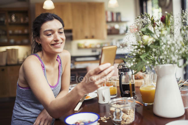 Lächelnde junge Frau beim SMS-Schreiben mit Smartphone am Frühstückstisch — Stockfoto