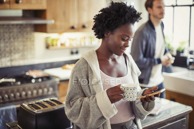 Donna che beve caffè, sms con smartphone in cucina — Foto stock