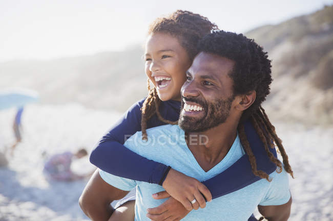Laughing pai piggybacking filha na praia ensolarada verão — Fotografia de Stock