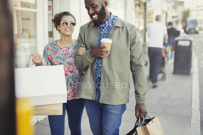 Lächelndes junges Paar mit Kaffee und Einkaufstüten, die an der Schaufensterfront entlanglaufen — Stockfoto