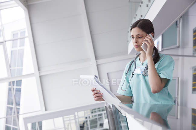 Female nurse with clipboard talking on cell phone in hospital — Stock Photo
