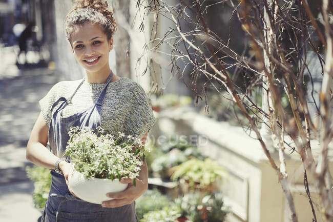Portrait de fleuriste souriante tenant une plante en pot à la devanture ensoleillée — Photo de stock