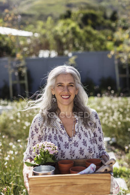 Retrato sorrindo mulher madura carregando bandeja de jardinagem no jardim ensolarado — Fotografia de Stock