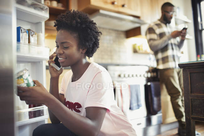 Woman talking on smart phone, reading label on jar in refrigerator in kitchen — Stock Photo