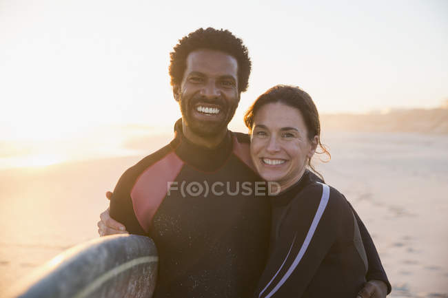 Retrato sorrindo, casal multi-étnico confiante com prancha de surf na ensolarada praia de verão ao pôr do sol — Fotografia de Stock