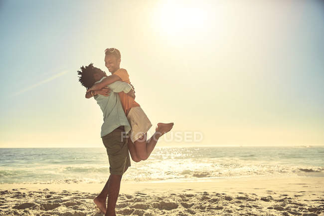 Petit ami ludique soulevant petite amie sur la plage ensoleillée de l'océan d'été — Photo de stock