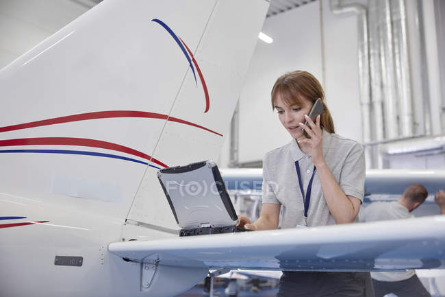 Female airplane engineer working at laptop and talking on cell phone in hangar — Stock Photo