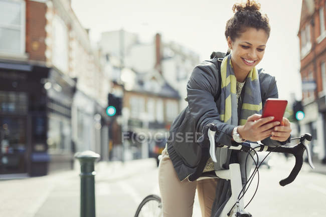 Giovane donna sorridente pendolarismo con la bicicletta, sms con il cellulare sulla strada urbana soleggiata — Foto stock