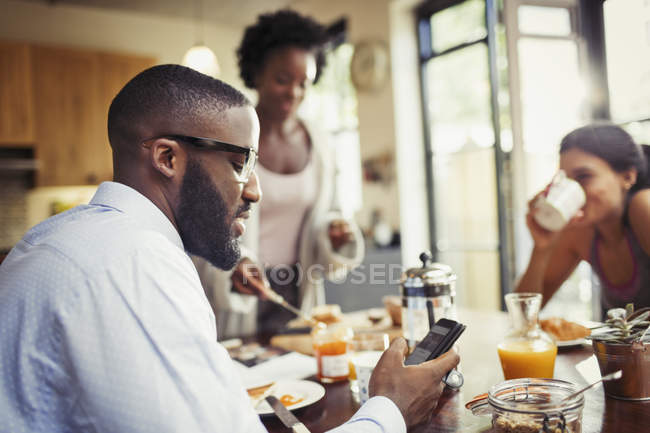 Hombre bebiendo café y mensajes de texto con teléfono inteligente en la mesa del desayuno - foto de stock