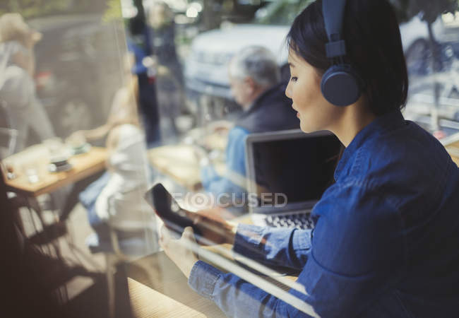 Young woman listening to music with headphones, texting with cell phone at cafe window — Stock Photo