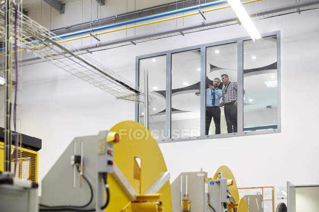 Male supervisors talking at window above factory floor — Stock Photo