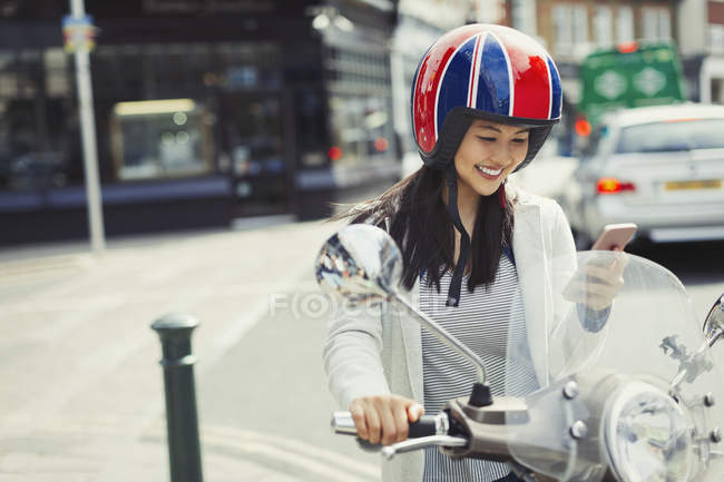 Smiling young woman texting with cell phone on motor scooter, wearing helmet on urban street — Stock Photo