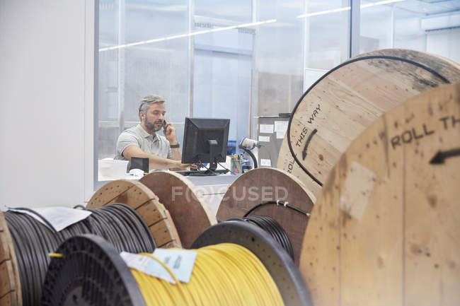Supervisor masculino trabajando en la computadora y hablando en el teléfono ell detrás de carretes en la fábrica de fibra óptica - foto de stock