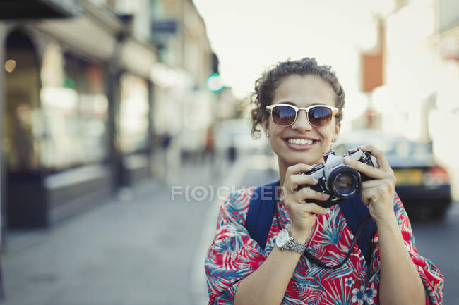Portrait smiling young female tourist in sunglasses photographing with camera on urban street — Stock Photo