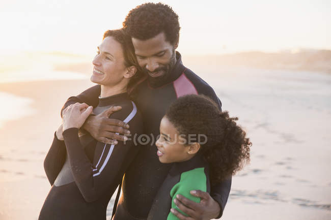 Ласковая многонациональная семья в мокрых костюмах, обнимающаяся на пляже на закате — стоковое фото