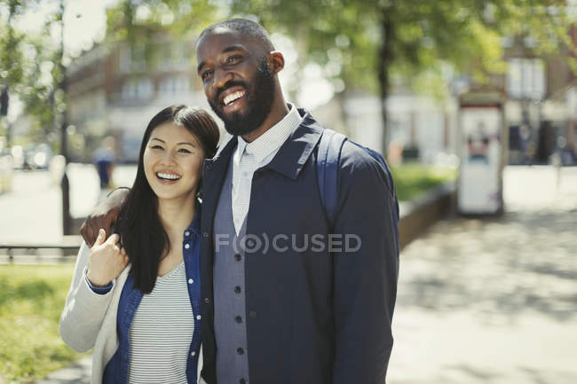 Sonriente y cariñosa pareja joven abrazándose en el soleado parque urbano - foto de stock