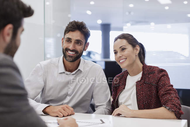 Улыбающаяся пара клиентов разговаривает с продавцом автомобилей в офисе автосалона — стоковое фото