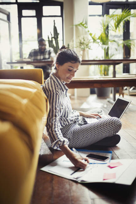 Femme pigiste travaillant à un ordinateur portable, prenant des notes sur le sol du salon — Photo de stock