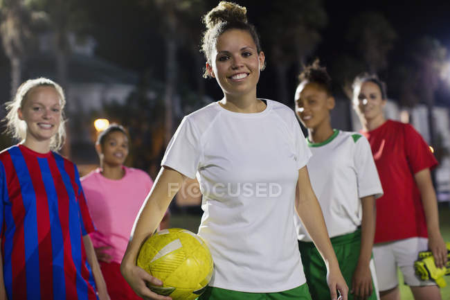 Retrato sonriente, confiado equipo de fútbol femenino joven con pelota en el campo por la noche - foto de stock