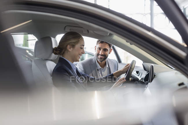 Vendedor de coches mostrando coche nuevo a cliente femenino en el asiento del conductor en la sala de exposición de concesionarios de automóviles - foto de stock