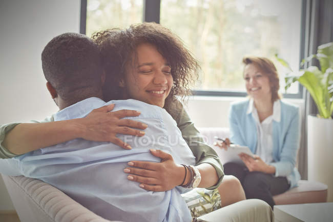 Sonriente terapeuta femenina viendo a la pareja abrazándose en la sesión de terapia de parejas - foto de stock