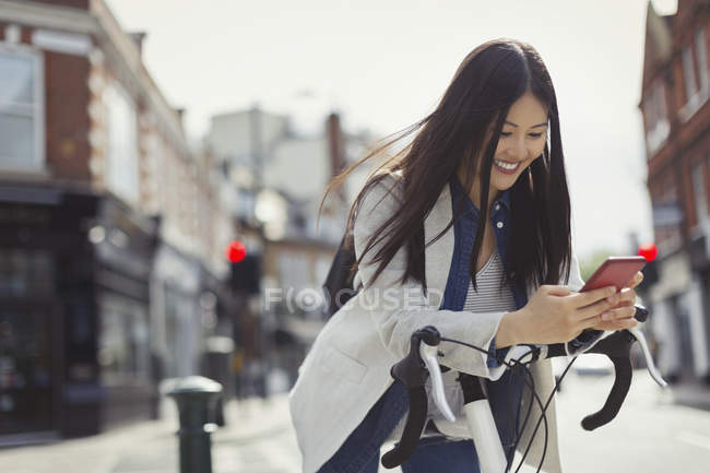 Улыбающаяся молодая женщина, путешествующая на велосипеде, переписывающаяся с сотовым на солнечной городской улице — стоковое фото