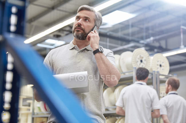 Superviseur masculin avec presse-papiers parlant sur le téléphone cellulaire dans l'usine de fibre optique — Photo de stock