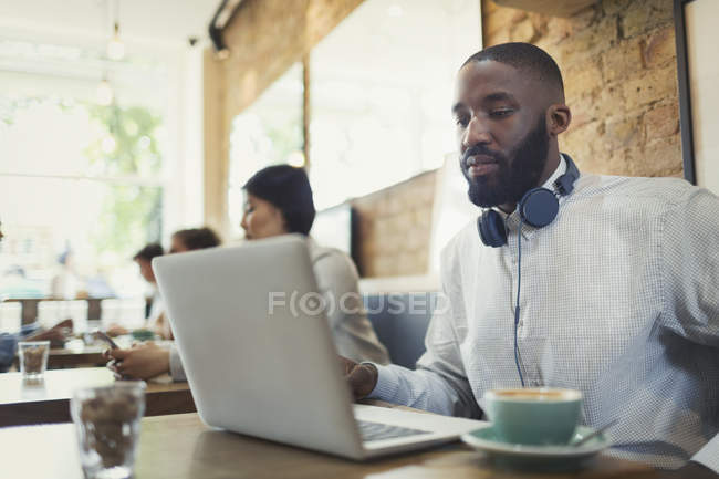 Young man with headphones using laptop and drinking coffee in cafe — Stock Photo