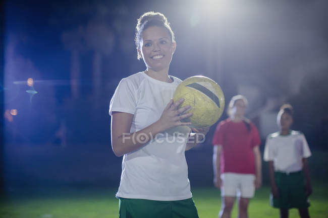 Retrato sorridente, confiante jovem jogadora de futebol feminina segurando bola de futebol em campo à noite — Fotografia de Stock