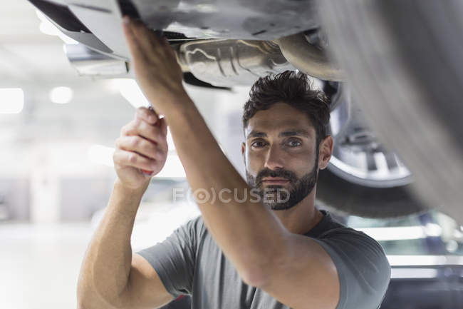 Retrato mecánico masculino serio que trabaja debajo del coche en taller de reparación de automóviles - foto de stock