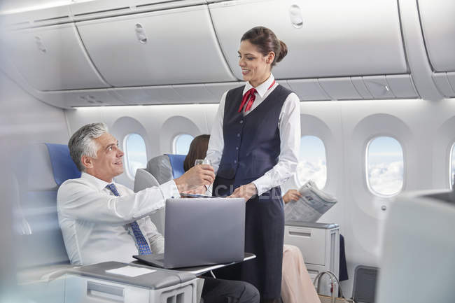 Assistente de bordo servindo bebida para homem de negócios que trabalha no laptop no avião — Fotografia de Stock