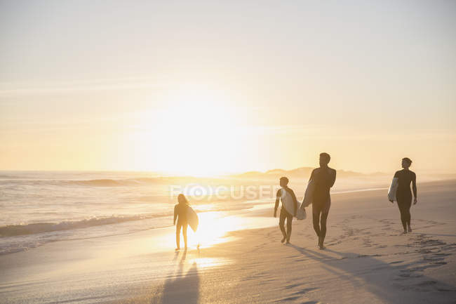 Surfistas de la familia Silhouette caminando con tablas de surf en la idílica y soleada playa de atardecer de verano - foto de stock