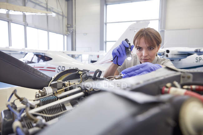Focused ingegnere meccanico femminile con torcia elettrica esaminando motore aereo in hangar — Foto stock