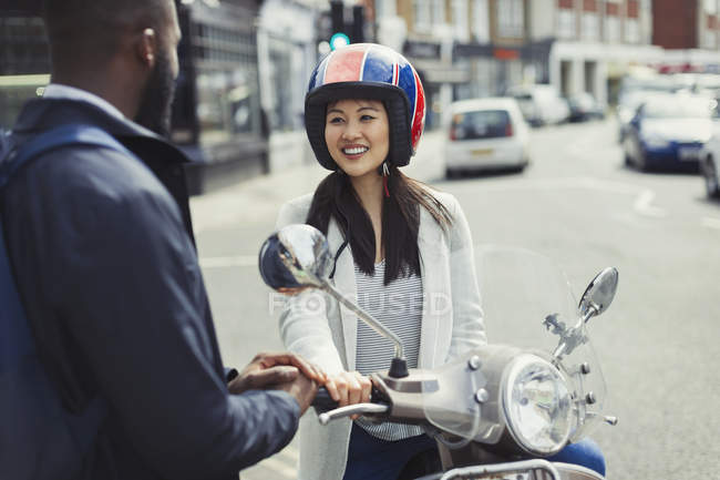 Souriant jeune femme sur scooter moteur parler à un ami sur la rue urbaine ensoleillée — Photo de stock