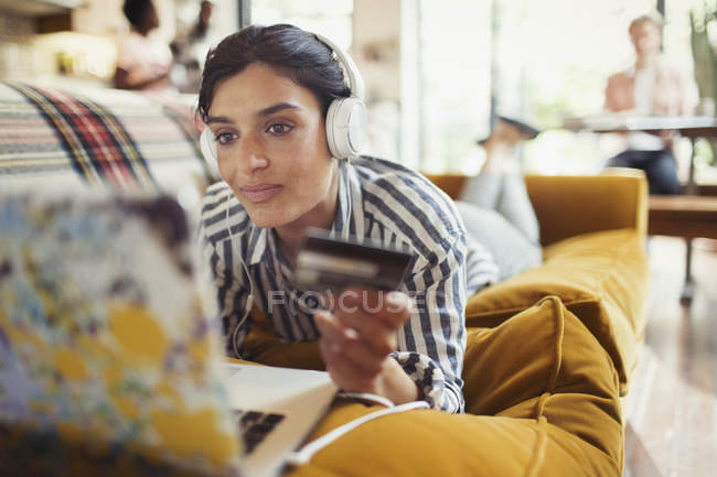 Mujer joven con tarjeta de crédito y auriculares compras en línea en el ordenador portátil en el sofá de la sala de estar - foto de stock