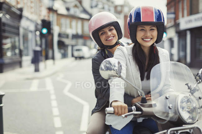 Smiling young women friends wearing helmets, riding motor scooter on urban street — Stock Photo