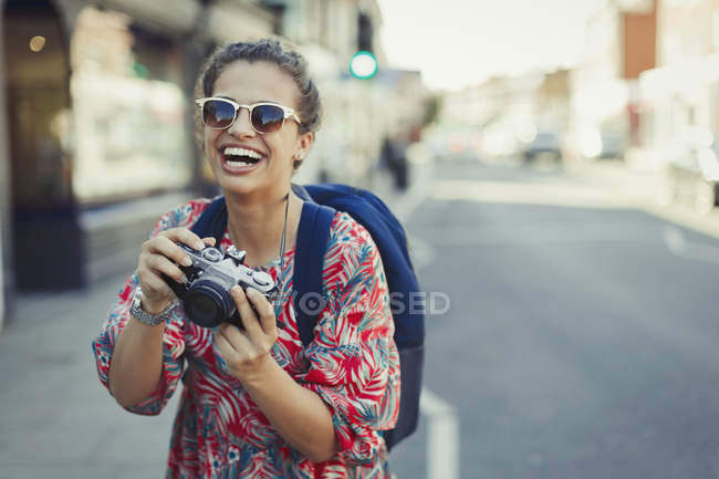 Retrato risueño, joven turista entusiasta en gafas de sol fotografiando con cámara en calle urbana - foto de stock