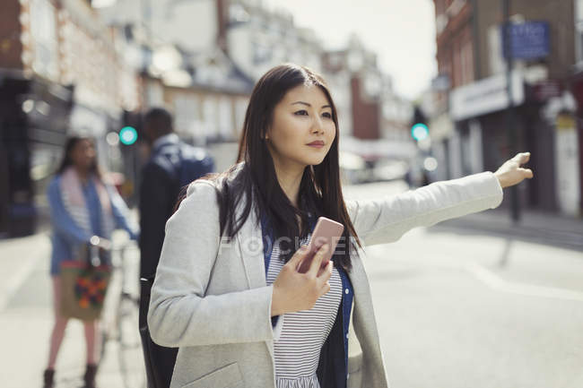 Junge Geschäftsfrau mit Handy bejubelt Taxi auf sonniger Stadtstraße — Stockfoto