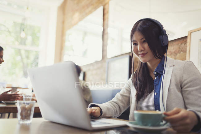 Sorrindo jovem ouvindo música com fones de ouvido no laptop e bebendo café no café — Fotografia de Stock