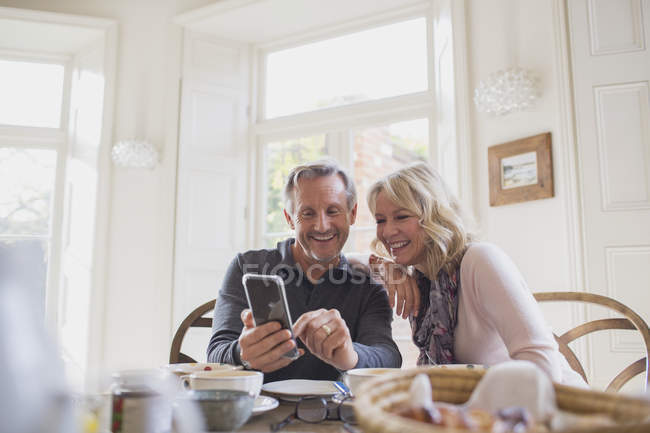 Усміхнена зріла пара використовує смартфон за обіднім столом — стокове фото