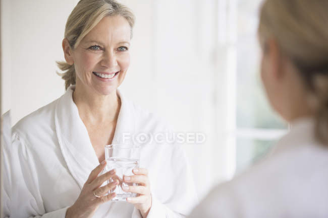 Усміхнена, впевнена зріла жінка в халаті п'є воду у дзеркалі ванної кімнати — стокове фото